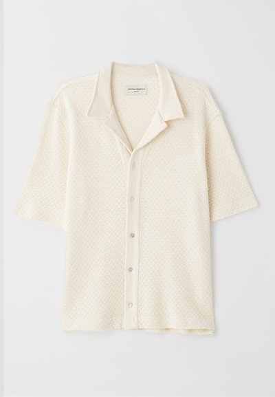 Kreminės spalvos trumpomis rankovėmis priekyje su apykakle, tekstūruotas audinys su deimantų raštais, penkių sagų užsegimas ir laisvas siluetas.