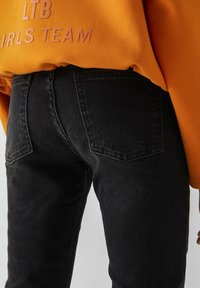 Černé džíny s úzkým střihem, které mají klasický pětikusový styl a viditelné švy. Spárované s oranžovou mikinou.