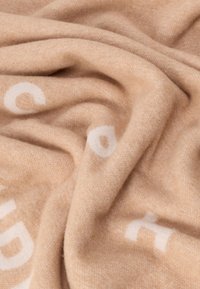 Beige tæppe lavet af blødt stof med hvid tekst i en kontrasterende skrifttype, med en glat, struktureret overflade. Krøllet og lagdelt.