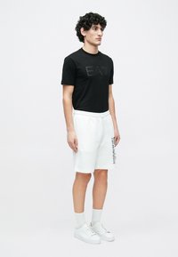 Νεαρός άνδρας στέκεται φορώντας μαύρο T-shirt EA7, λευκές βερμούδες Emporio Armani, λευκές κάλτσες και λευκά αθλητικά παπούτσια, μπροστά σε απλό φόντο.