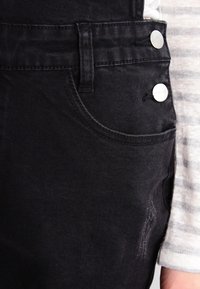 Black denim overalls with a pocket, featuring silver button accents and a distressed area. Background shows a gray and white striped shirt.