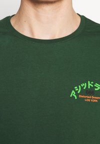 Tmavě zelené bavlněné tričko s kulatým výstřihem a krátkým rukávem. Na přední straně se nachází text v zelené, Oranžové a černé barvě s grafickým designem.