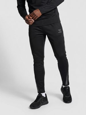 Man draagt zwarte slim-fit sportbroek, zwart langarmshirt, zwarte sneakers en witte sokken en staat tegen een effen achtergrond.