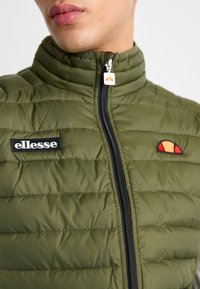 Jako prošiveno maslinasto zelene boje s visokim ovratnikom, crnim zatvaračem, crnim logom "ellesse" na zakrpli i šarenim grbom blizu ramena.