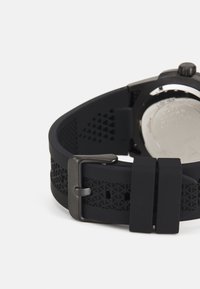 Correa de reloj de goma negra con recortes geométricos, herrajes plateados y una hebilla negra mate, que presenta una superficie texturizada suave.