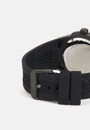 Schwarzes Gummiband mit geometrischen Ausschnitten, silbernen Beschlägen und einer mattschwarzen Schnalle, die eine glatte, strukturierte Oberfläche aufweist.