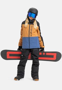 Person mit Skihelm und Skibrille, der ein Snowboard mit rotem Design hält, trägt eine farblich abgesetzte Jacke und schwarze Schneehosen vor weißem Hintergrund.