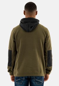 Olivgrüner Fleece-Hoodie mit schwarzer Kapuze und schwarzen Patches an den Ärmeln. Weicher Stoff, lässige Passform und gerippte Bündchen.