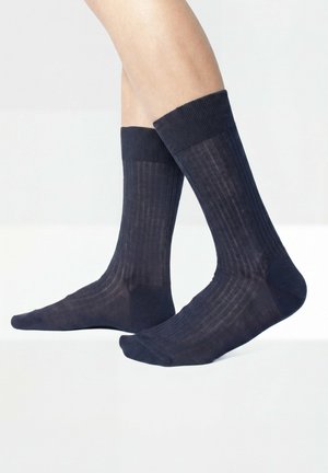 Beine mit dunkelblauen, gerippten Socken bis zur Mitte der Wade mit verstärkten Zehen und Fersen vor einem schlichten hellen Hintergrund.