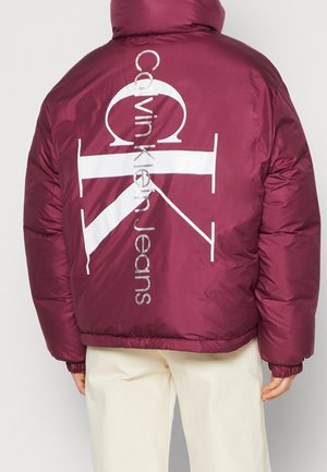 Personne portant une doudoune bordeaux avec un grand logo blanc "Calvin Klein Jeans" dans le dos et un pantalon blanc sur un fond uni.
