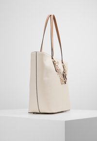 Beżowa skórzana torba tote z dwoma brązowymi uchwytami, z dekoracyjnymi metalowymi akcentami, teksturowaną powierzchnią i usztywnionym kształtem.