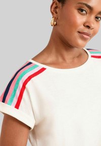 Kreminės spalvos marškiniai su suktomis rankovėmis, ant pečių puošti daugiaspalvėmis juostelėmis raudonos, rožinės, melsvai žalios ir tamsiai mėlynos spalvų. Slydžios tekstūros.