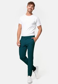 Pantalons turquoise en tissu texturé, associés à un t-shirt blanc uni. Présentent une coupe ajustée et des poches, portés avec des baskets blanches.
