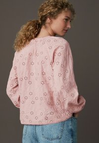 Blusa rosa ricamata con ritagli floreali, scollatura rotonda e maniche a tre quarti. Abbinata a jeans in denim blu chiaro.