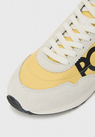 Detalizēts skats uz sporta apaviem ar dzeltenu auduma virsu, pelēkiem zamša akcentiem un baltām šņorēm, ar melnu logotipu sānos.