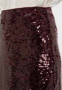 Jupe en sequins bordeaux avec une texture lisse et scintillante, composée de sequins circulaires superposés qui reflètent la lumière et créent une profondeur visuelle.