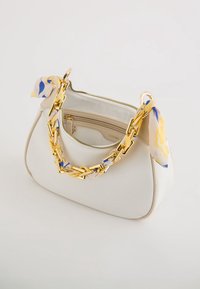 Bílá kabelka se zlatým řetízkovým uchem, obtočeným žlutým a modrým vzorovaným šátkem, otevřený zip odhalující béžovou vnitřní kapsu.