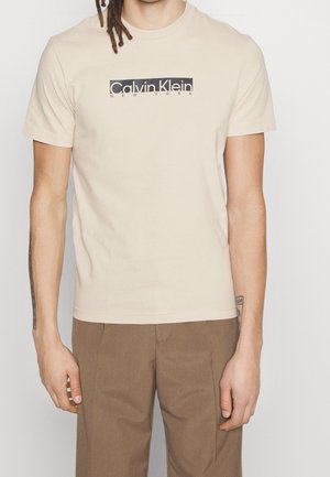 Homme portant un t-shirt beige Calvin Klein New York et un pantalon marron, debout devant un fond clair uni.