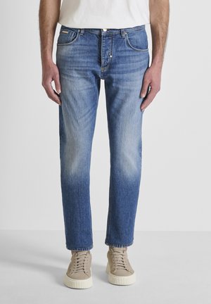 Hombre con vaqueros desgastados azul claro, zapatillas deportivas beige y una camisa blanca, posando frente a un fondo liso de color gris claro.