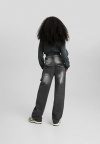 Kvinna med lockigt hår som bär bleknade svarta cargobyxor och en svart sweatshirt, ståendes med ryggen mot en simpel grå bakgrund.