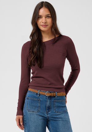 Jeune femme aux longs cheveux foncés portant un haut ajusté à manches longues bordeaux, un jean bleu avec des poches avant et une ceinture marron, debout devant un fond uni.