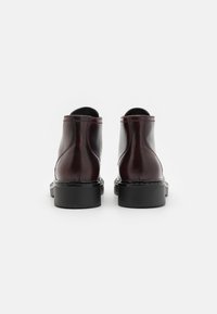 Botas de tobillo de cuero burdeos con un acabado suave, suela de goma negra y detalles cosidos a lo largo de lascosturas, vistas desde la parte posterior.