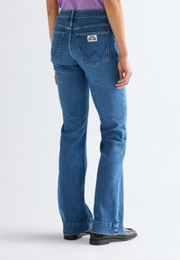 Personne portant un jean bootcut bleu avec poches arrière et écusson logo, associé à des chaussures noires et un haut violet, debout devant un fond blanc uni.