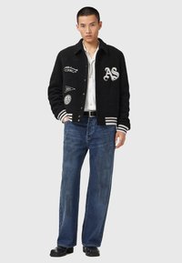 Ung mand iført en sort varsityjakke med patches, hvid skjorte, blå jeans og sorte sko, stående foran en ensfarvet grå baggrund.