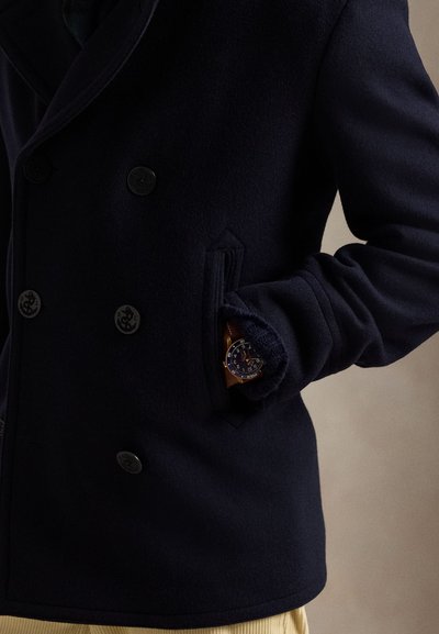 Person iført en mørk marineblå dobbeltradet jakke med ankerknapper og beige bukser, hånd i jakkelommen, der viser et armbåndsur.