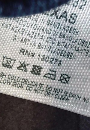 Gondozási címke, amely a 4S méretet mutatja, Bangladesben készült, mosási szimbólumok, fehérítés nélküli, alacsony hőfokon vasalható, vegytisztítás tilos, és szövetápolási utasítások.