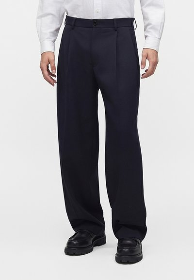 Homme portant un pantalon noir à jambes larges, des chaussures noires et une chemise blanche à manches longues boutonnée, debout sur un fond blanc uni.