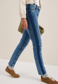 Blå denimjeans med rak passform, ljusare tonade sidosträck och en klassisk femficksdesign. Bärs med bruna sneakers.