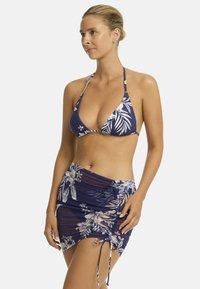 Modelo vistiendo un top de bikini floral en tonos azul marino y una falda arrugada a juego, de pie con un ligero perfil, mostrando atuendo de playa.