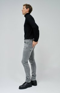 Suéter negro de cuello alto, jeans gris ajustados y zapatos negros con cordones. Tejido suave con detalles sutiles de desgaste en los jeans. Vista lateral de pie.