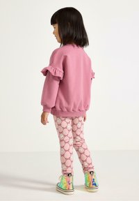 Vaate yhdistelmä, jossa on vaaleanpunainen huppari, jonka hihansuissa on röyhelöitä, yhdistettynä vaaleanpunaisiin leggingseihin, joissa on toistuva leijonakuosi. Heijastavat, moniväriset korkeavartiset tennarit.
