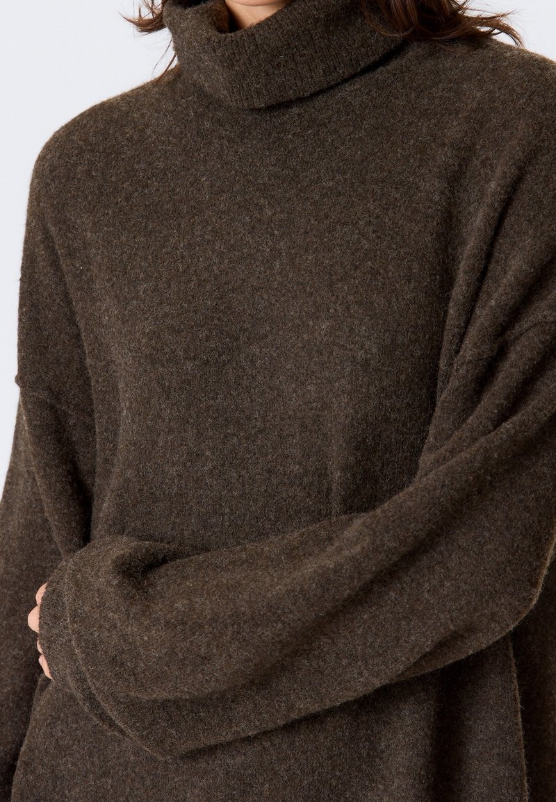 Pull col roulé en laine marron doux avec de longues manches amples porté par une personne croisant les bras sur le torse.