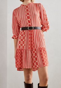 Robe à motifs rouge et crème avec un devant à boutons, une taille froncée et des manches trois quarts. Finition avec une ceinture noire et des bottes jusqu'aux genoux.