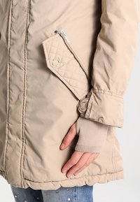 Giacca beige con tasca trapuntata e zip, dettaglio a bottone sui polsini e apertura maniche a coste. Tessuto liscio e leggermente lucido.