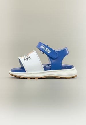 Children’s sandal with blue straps, white front band featuring a teddy bear, and white cushioned sole with brown tread.