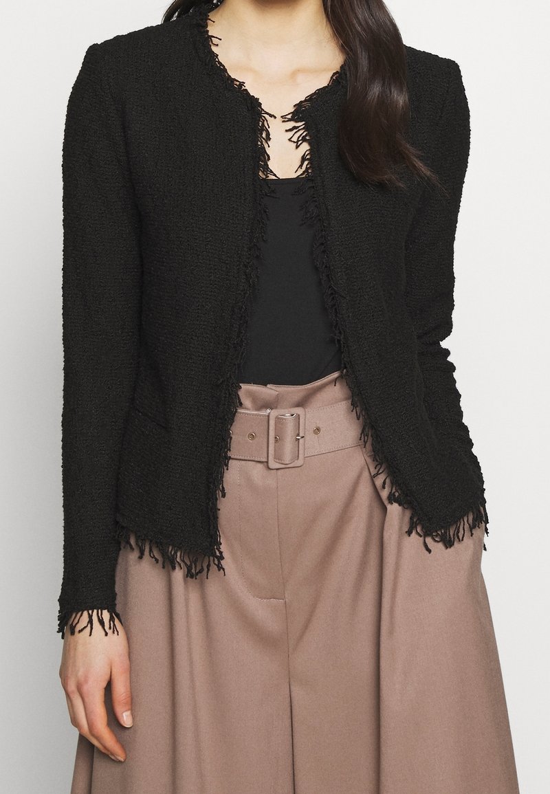 Femme portant une veste noire texturée avec des bords frangés sur un haut noir, associée à un pantalon large taille haute couleur taupe avec une ceinture.