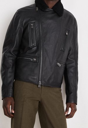 Homme portant une veste en cuir noire avec plusieurs poches zippées et un pantalon vert olive foncé sur un fond clair uni.