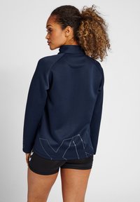 Marinblå atletisk jacka med hög krage, långa ärmar och ett geometriskt vitt accentdesign på nedre delen av ryggen. Materialet verkar slätt.