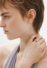 Anillo de oro con una gema púrpura central, llevado en una mano que descansa sobre el cuello, con pelo corto y un atuendo gris en vista de perfil.