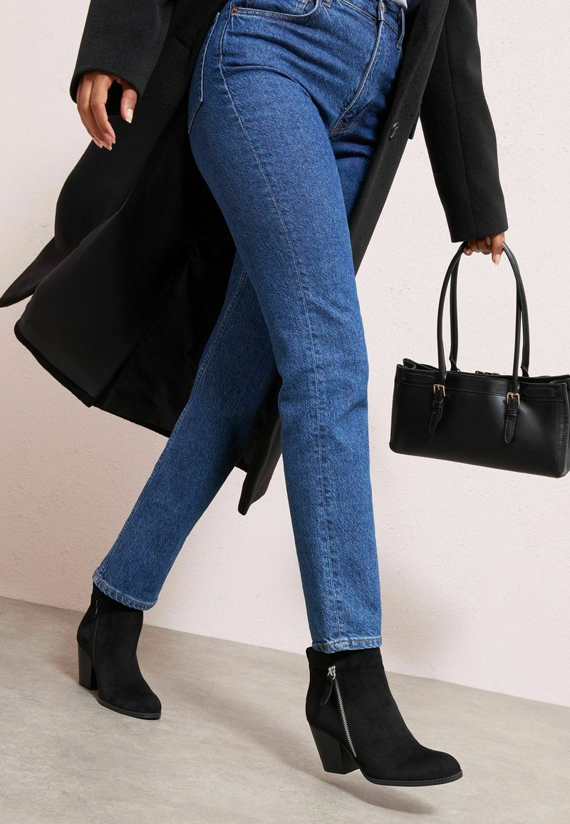 Botines negros de ante hasta el tobillo con un detalle de cremallera, jeans azules y un bolso negro. La persona está caminando, llevando un abrigo largo negro.