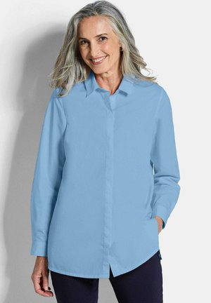 Femme aux cheveux gris portant une chemise à manches longues bleu clair boutonnée et un pantalon foncé, souriante, avec une main dans la poche.