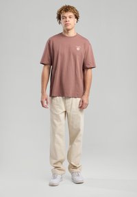 Ruskea lyhytnhihainen t-paita, jossa on pieni kukkakuvio, yhdistettynä vaaleanbeigeihin löysiin housuihin ja valkoisiin lenkkareihin. Yksinkertainen, rento asuste.