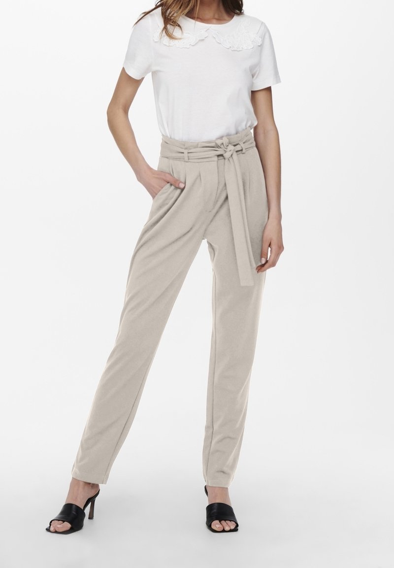 Pantalon beige taille haute avec une ceinture à la taille, des plis à l'avant et des poches latérales. Associé à un haut blanc à manches courtes et des talons noirs.