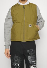 Olivgrön quiltad väst med grå ärmar, blixtlås framtill, två framfickor och en logotyp-patch på vänster sida.