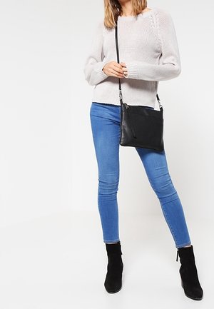 Kobieta ubrana w beżowy sweter, niebieskie obcisłe dżinsy, czarne botki za kostkę, niosąca czarną torebkę na ramię z detalem zamka.