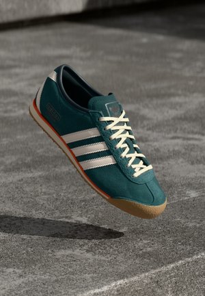 Turkis Adidas sneaker med hvide striber og snørebånd, orange kant og gummisål, svævende over en grå betonoverflade.